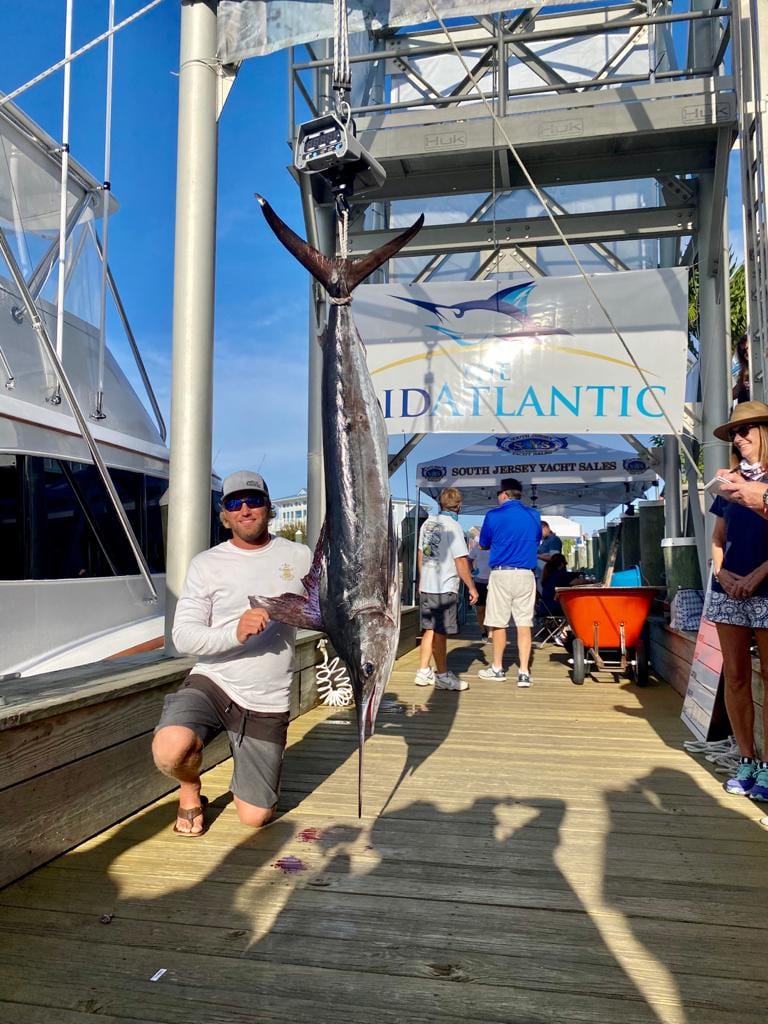 Zak Smith kneeling beside a white marlin hanging from the scale under The MidAtlantic banner