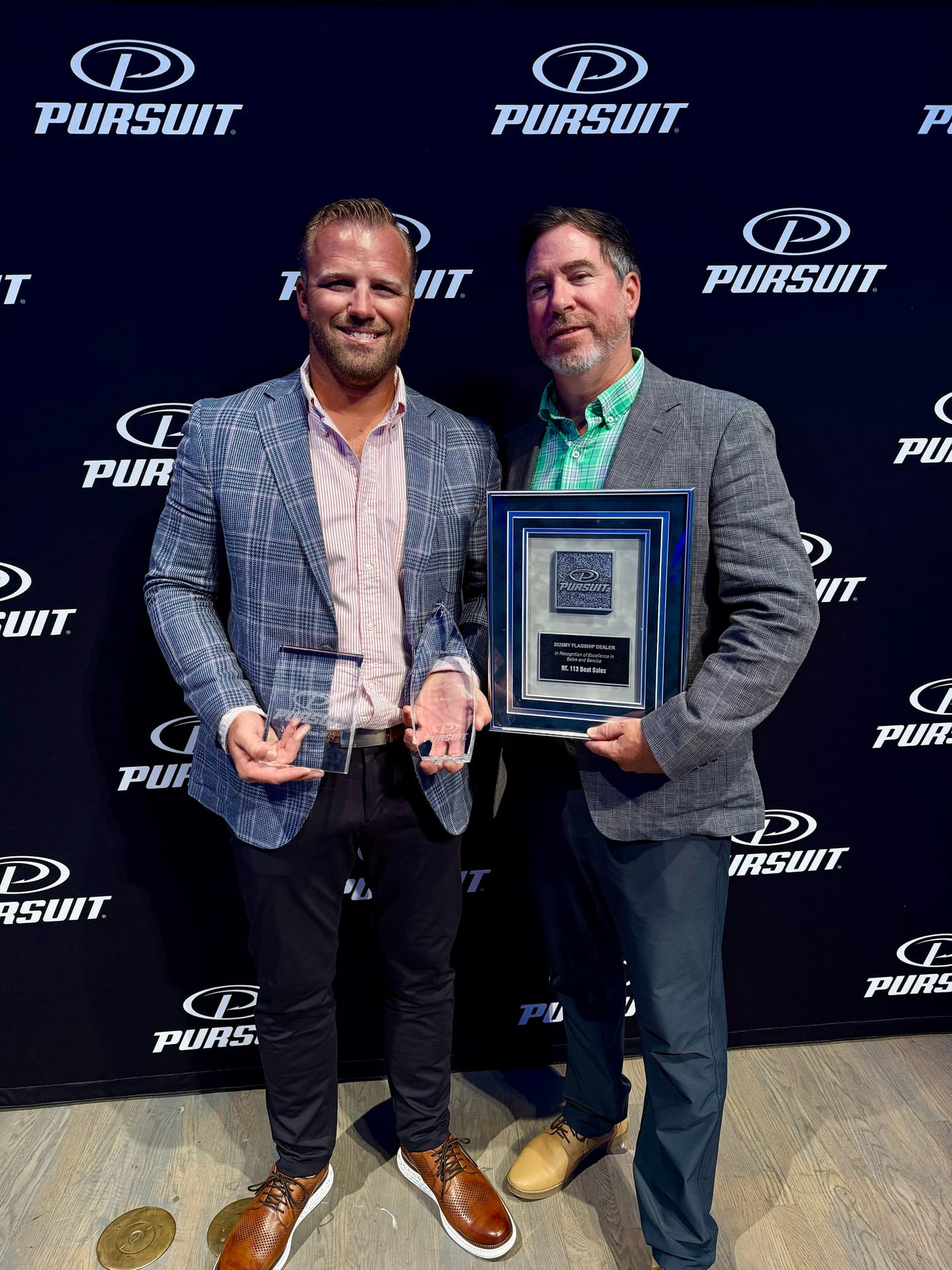 Zak Smith holding two glass trophies beside a Pursuit representative holding the 2025 Flagship Dealer plaque, in front of a Pursuit backdrop