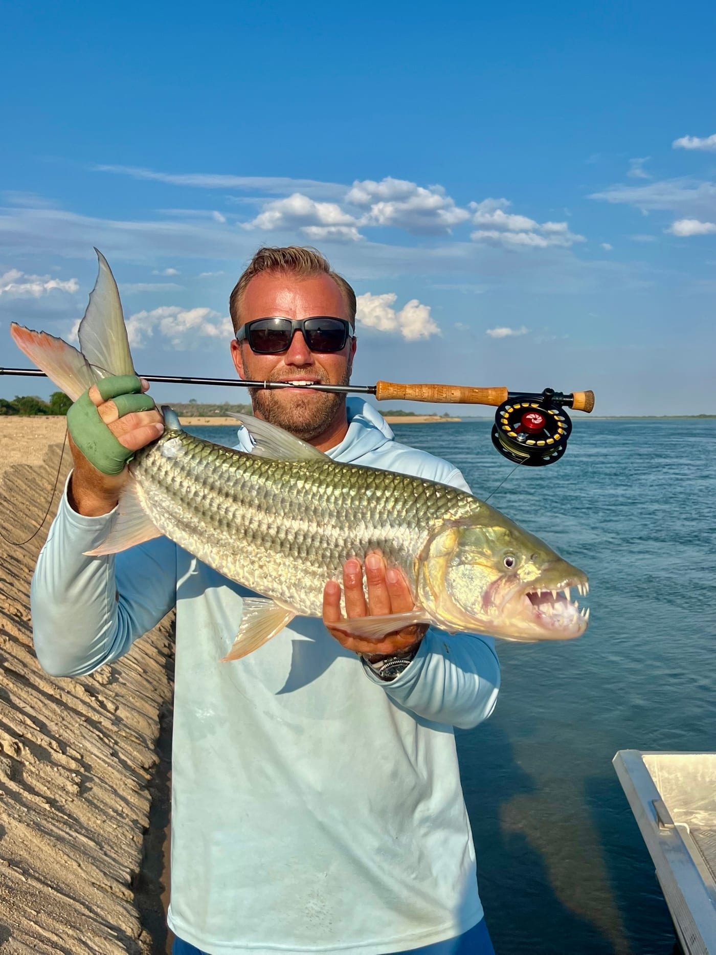 Zak Smith holding a tigerfish with a fly rod gripped in his teeth, an African river behind him